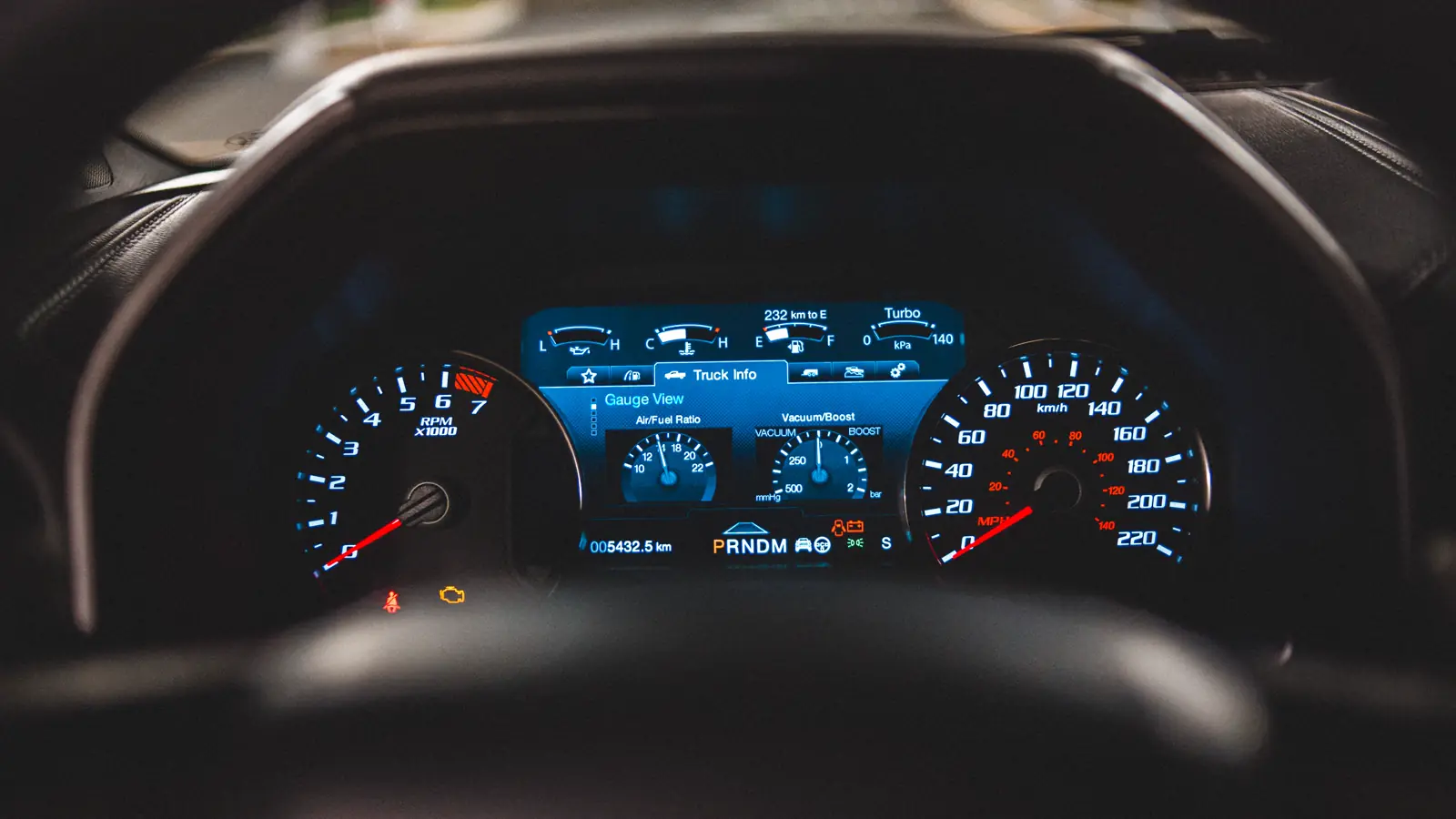 Yellow check engine light illuminated on a vehicle dashboard viewed through a steering wheel